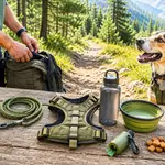 Dog hiking gear laid out on a trailside picnic table: a collar with an ID tag, a rope leash, a harness, a water bottle, a collapsible bowl, a waste-bag dispenser and treats, with a dog waiting beside them.
